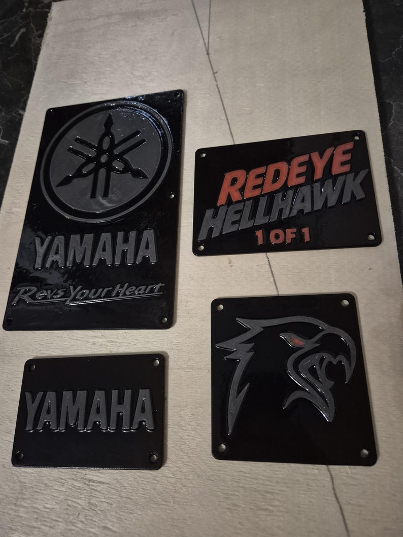 May include: Four black metal plaques with embossed designs. One plaque features the Yamaha logo and text, another reads "Redeye Hellhawk 1 of 1", and the others have stylized graphics. All plaques have mounting holes.