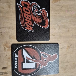 May include: Two black rectangular cards with textured surfaces. One card features a red cobra logo with white outlines and the word "COBRA". The other card has a red and white logo with the word "AEON" and a silhouette of a person.