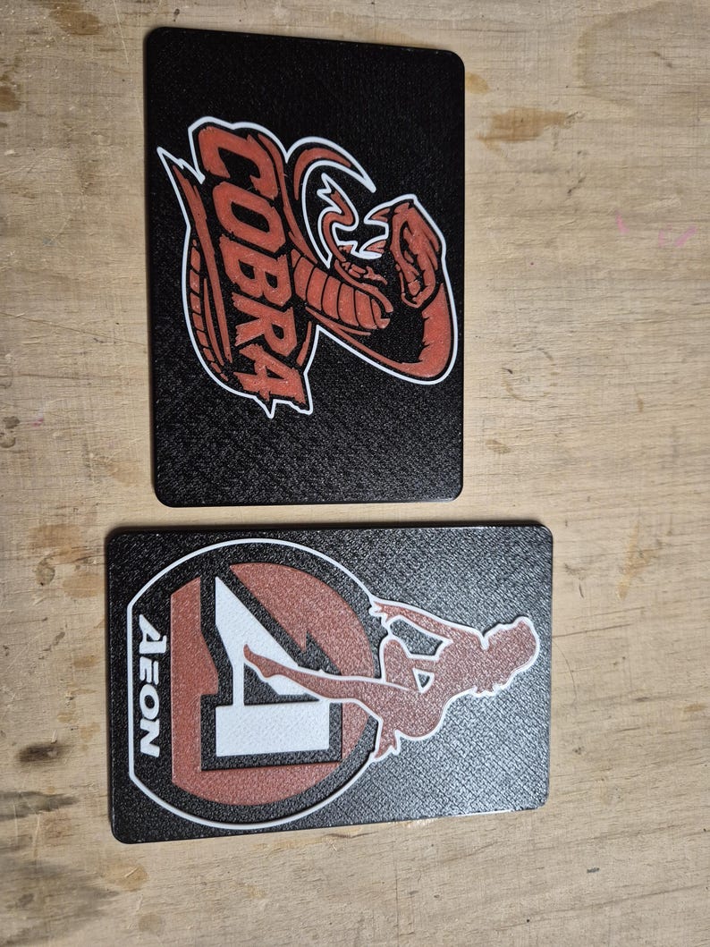 May include: Two black rectangular cards with textured surfaces. One card features a red cobra logo with white outlines and the word "COBRA". The other card has a red and white logo with the word "AEON" and a silhouette of a person.