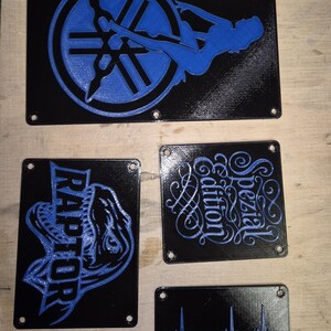 May include: A set of black rectangular plaques with blue designs. One plaque shows a Yamaha logo and a silhouette. Another says "RAPTOR". Other plaques have the words "Spezial Edition" and a heartbeat with a scorpion.