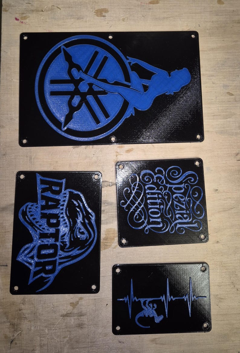 May include: A set of black rectangular plaques with blue designs. One plaque shows a Yamaha logo and a silhouette. Another says "RAPTOR". Other plaques have the words "Spezial Edition" and a heartbeat with a scorpion.
