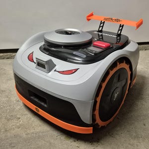 Könnte beinhalten: Ein Roboter-Rasenmäher mit grau-schwarzem Gehäuse, orangefarbenen Akzenten und einem Heckspoiler. Der Mäher hat eine kreisförmige Oberseite, ein digitales Display und ein lächelndes Gesichtsdesign mit roten Akzenten. Die Räder sind schwarz mit orangefarbener Zierleiste.