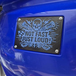 May include: A black rectangular plate with the text "NOT FAST JUST LOUD!" in blue, featuring a skull and sound wave graphics. The plate is fixed to a blue surface with four silver screws.