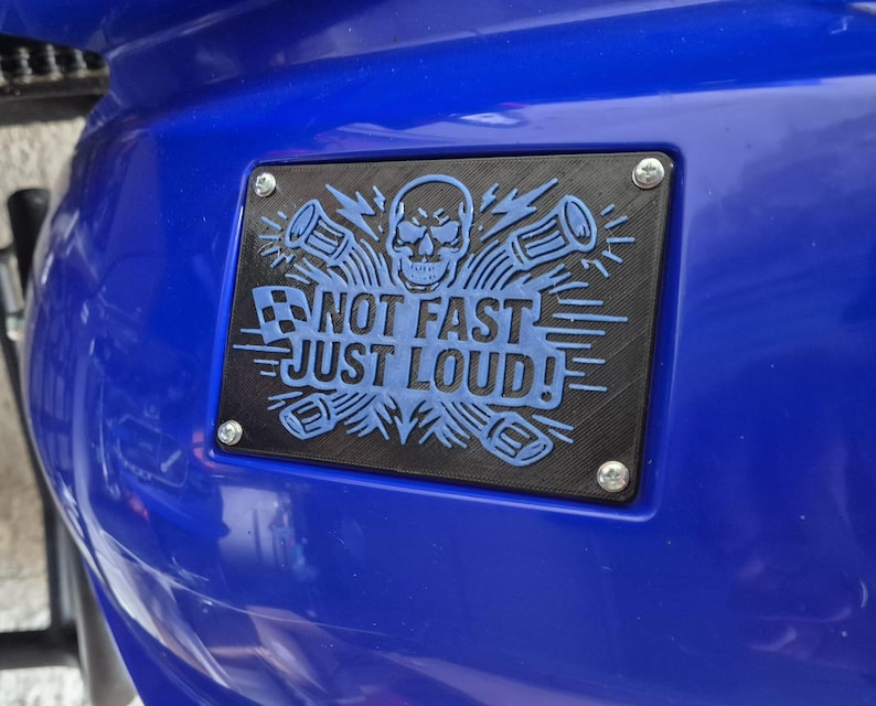 May include: A black rectangular plate with the text "NOT FAST JUST LOUD!" in blue, featuring a skull and sound wave graphics. The plate is fixed to a blue surface with four silver screws.