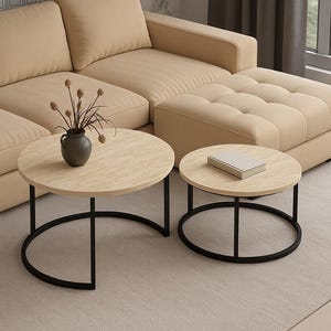 Puede incluir: Dos mesas de centro redondas con tapas de madera clara y bases de metal negro. Una mesa es más grande, con una base curva, y la otra es más pequeña con una base circular. Un sofá y un otomano beige están en el fondo.