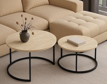 Puede incluir: Dos mesas de centro redondas con tapas de madera clara y bases de metal negro. Una mesa es más grande, con una base curva, y la otra es más pequeña con una base circular. Un sofá y un otomano beige están en el fondo.
