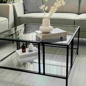 May include: A modern living room scene featuring a square glass coffee table with a black metal frame. The table holds decorative items, including a vase with dried flowers and books with the word "CEREAL" on the cover. A gray sofa and patterned pillow are in the background.