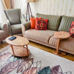 Könnte beinhalten: Wohnzimmer mit beigem Sofa, olivgrünen Kissen und dekorativen Kissen. Zwei Holztische und ein Teppich mit Blattmustern befinden sich vor dem Sofa. Ein grauer Sessel und eine Stehlampe sind ebenfalls zu sehen.