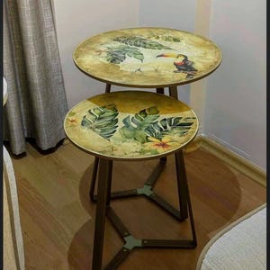 Peut inclure: Deux tables d'appoint rondes avec un thème tropical. Les plateaux de table présentent un toucan et des feuilles vertes luxuriantes sur un fond jaune. Les tables ont des pieds en métal brun foncé et sont imbriquées.