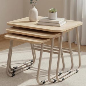 Puede incluir: Un juego de tres mesas nido con tapas de madera clara y patas de metal beige. Las mesas son cuadradas con esquinas redondeadas. Una pequeña suculenta en una maceta blanca y una pila de libros están sobre la mesa superior.