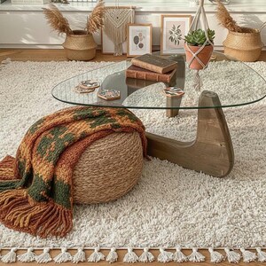 Könnte beinhalten: Wohnzimmerszene mit einem Couchtisch aus Glas mit Holzfuß, einem geflochtenen Ottoman mit einer Fransen-Decke und einem flauschigen weißen Teppich. Dekorative Elemente sind gerahmte Kunstwerke, eine Hängepflanze und Weidenkörbe.