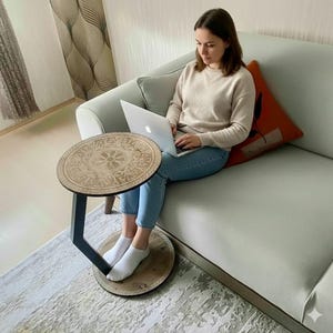 Pode incluir: Uma mesa lateral redonda de madeira com um design floral decorativo no tampo. A mesa tem um suporte de metal preto e uma base circular. A mesa está ao lado de uma pessoa sentada em um sofá verde claro.