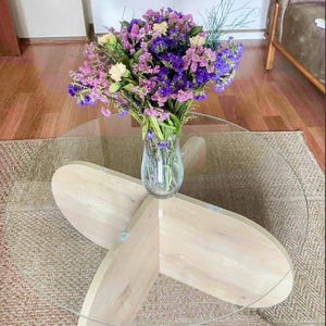 Könnte beinhalten: Ein runder Couchtisch mit Glasplatte und hellem Holzfuß, auf dem eine klare Vase mit lila, rosa und gelben Blumen steht. Der Tisch steht auf einem gewebten Teppich.