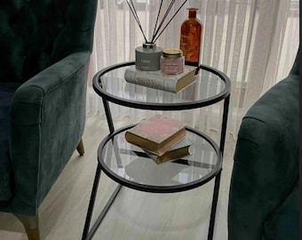 Puede incluir: Una mesa auxiliar de dos niveles con estructura de metal negro y estantes de vidrio transparente. La balda superior contiene un difusor, una vela y una botella decorativa. Hay libros en ambas baldas.