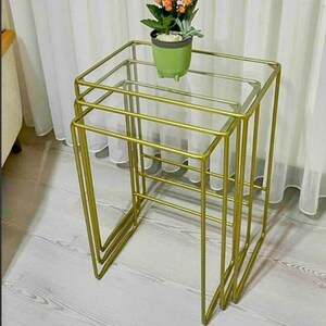 Puede incluir: Juego de tres mesas nido con estructura de metal dorado y tapas de cristal transparente. Las mesas están dispuestas en configuración anidada. Una pequeña maceta verde con flores naranjas se encuentra en la mesa superior. Decoración del hogar.