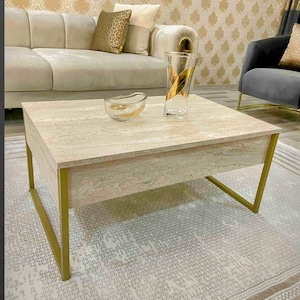May include: A square coffee table with a light beige stone top and gold-colored metal legs. The table has a modern design and is in a living room setting. A clear glass vase with gold accents and a small glass bowl are on the table.