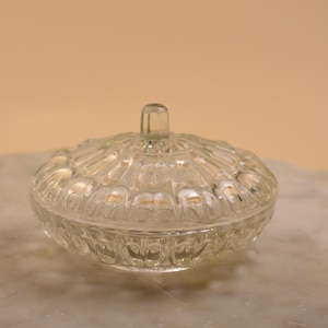 May include: A clear, vintage glass candy dish with a lid. The dish has a round shape with a textured, patterned design. The lid has a small handle. The dish is sitting on a marble surface.