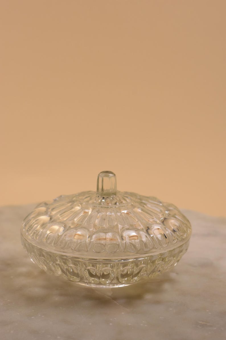 May include: A clear, vintage glass candy dish with a lid. The dish has a round shape with a textured, patterned design. The lid has a small handle. The dish is sitting on a marble surface.