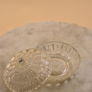 May include: A clear, vintage glass dish with a lid. The dish and lid feature a detailed, embossed pattern. The lid is slightly ajar, revealing the interior of the dish. The dish is round and sits on a marble surface.