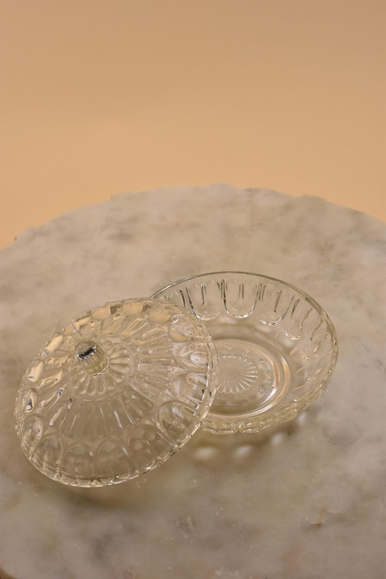 May include: A clear, vintage glass dish with a lid. The dish and lid feature a detailed, embossed pattern. The lid is slightly ajar, revealing the interior of the dish. The dish is round and sits on a marble surface.