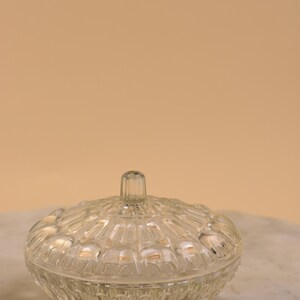 May include: A clear, vintage glass candy dish with a lid. The dish has a round shape with a textured, patterned design. The lid has a small, vertical handle. The dish is sitting on a marble surface.