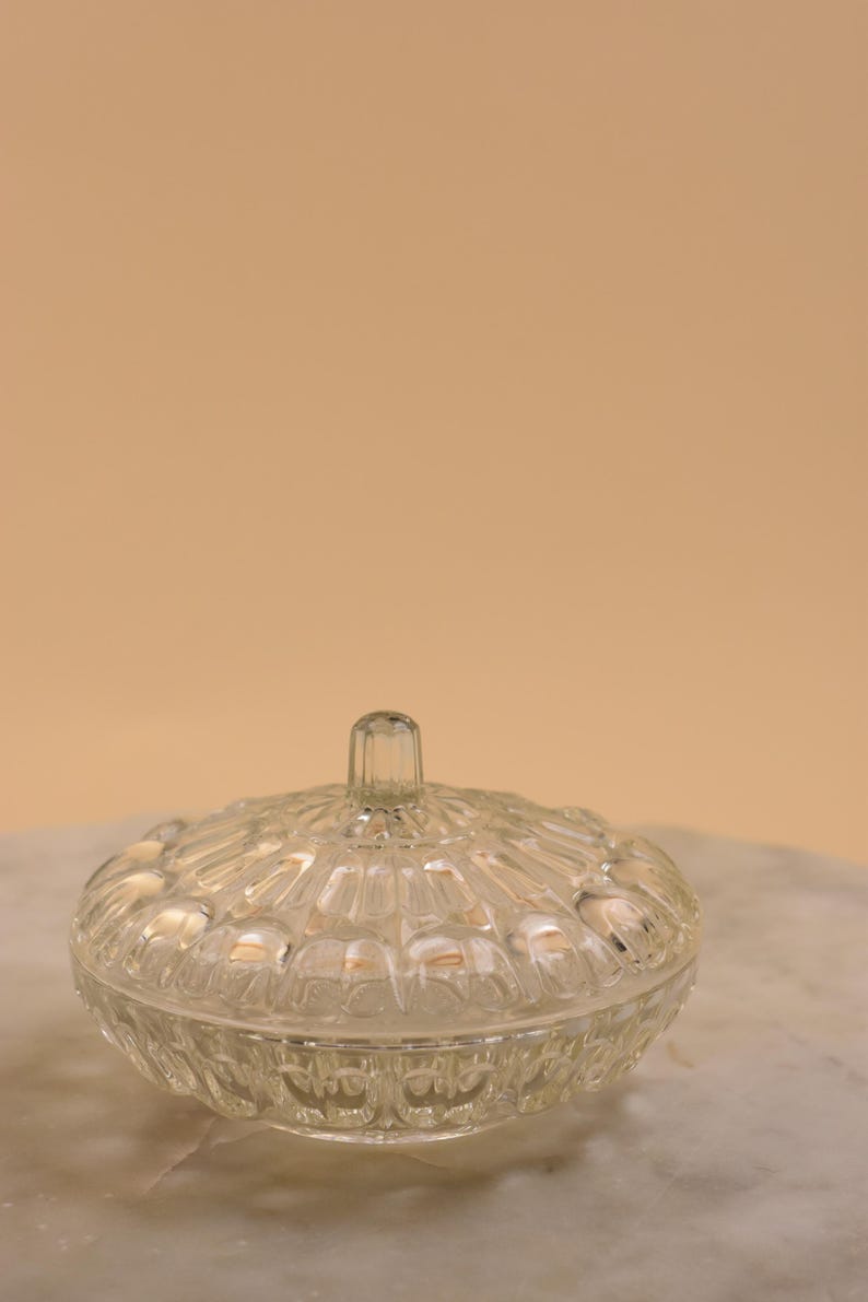 May include: A clear, vintage glass candy dish with a lid. The dish has a round shape with a textured, patterned design. The lid has a small, vertical handle. The dish is sitting on a marble surface.