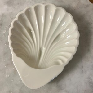 May include: A white ceramic seashell-shaped dish with a scalloped edge. The dish has a smooth, glossy finish and a small, curved handle. The interior features a ribbed design, adding texture to the decorative piece.