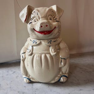 May include: A vintage ceramic pig-shaped cookie jar. The jar is off-white with red accents on the ears and mouth. It features blue floral details on the collar and overalls. The pig has a smiling expression.