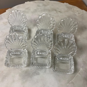 May include: Six clear glass seashell-shaped napkin rings. Each ring features a detailed fan-like design and a square base with a textured border. The napkin rings are arranged on a light-colored surface.