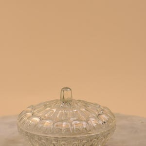 May include: A clear, lidded glass jar with a textured, rounded design. The lid has a small, rectangular handle. The jar sits on a marble surface against a neutral background. Ideal for storing small items.