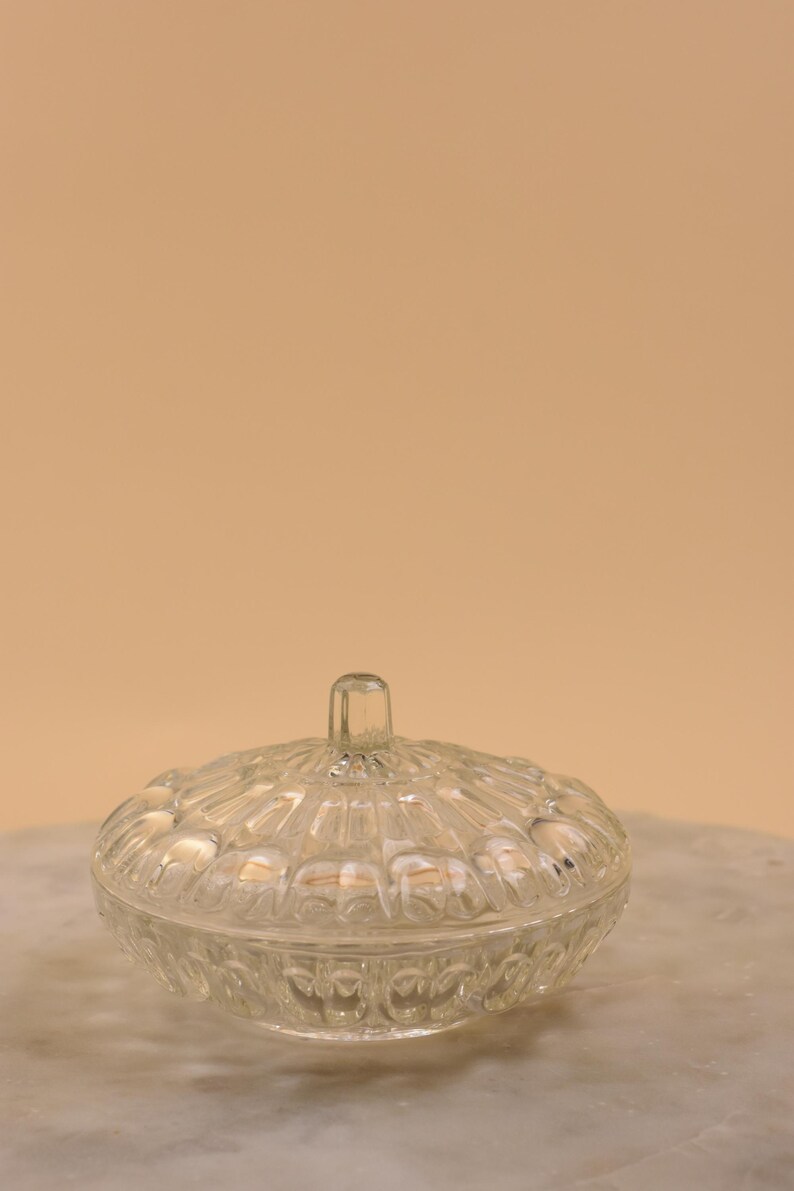 May include: A clear, lidded glass jar with a textured, rounded design. The lid has a small, rectangular handle. The jar sits on a marble surface against a neutral background. Ideal for storing small items.