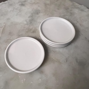 May include: A stack of four and one individual white ceramic coasters. The coasters are round with a slightly raised edge. The coasters are on a gray and white marble-like surface.