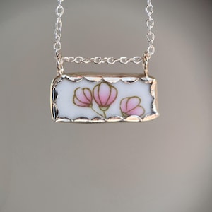 May include: A silver chain necklace with a rectangular pendant. The pendant features a white background with pink floral designs and a silver border. The necklace is a delicate piece of jewelry.