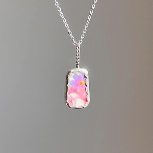 May include: A silver necklace with a rectangular pendant. The pendant features a pink and white background with a purple and yellow flower design. The chain is delicate and the pendant has a textured border.