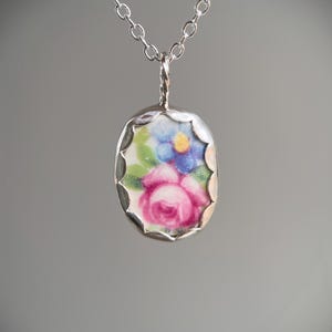 May include: A silver-toned necklace featuring a floral pendant. The pendant has a porcelain oval with a pink rose and blue flowers. The pendant is framed in a scalloped silver setting and hangs from a delicate chain.