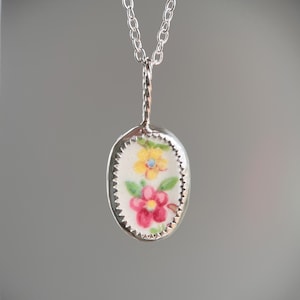 May include: A silver-toned pendant necklace featuring an oval-shaped pendant with a floral design. The pendant showcases a cluster of pink and yellow flowers with green leaves on a white background, framed by a scalloped edge. The chain is a delicate link style.