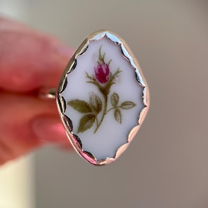May include: A gold-toned ring featuring a white, oval-shaped centerpiece with a hand-painted rose design. The rose has a deep pink bud, green leaves, and a delicate stem. The ring's setting has a scalloped edge.