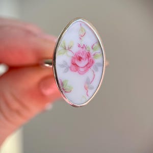May include: Oval-shaped ring with a silver band. The ring features a white porcelain inlay with a pink rose and green leaves design. The floral pattern is centered on the ring's face, creating a delicate and feminine aesthetic.