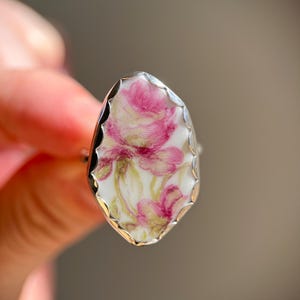 May include: A silver ring with a floral design. The ring features a white background with pink and yellow floral artwork. The ring has a scalloped edge and a polished silver band. The ring is a unique piece of jewelry.