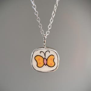 May include: A silver necklace featuring a pendant with a butterfly design. The butterfly has orange wings with black outlines and a pink body, set against a white background. The pendant is encased in a silver frame.