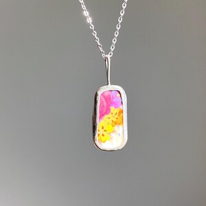 May include: A silver necklace with a rectangular pendant. The pendant features a colorful floral design with pink, yellow, and orange hues. The chain is delicate and the pendant is set in a silver frame.