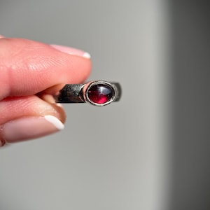 May include: A silver ring with a round, deep red gemstone. The ring's band has a textured, hammered appearance. The gemstone is set in a bezel setting, showcasing its rich color. The ring is held up against a soft gray background.