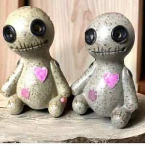 May include: Two gray voodoo dolls with black button eyes and stitched mouths. Each doll has a pink heart and patch. They sit on a wooden base with a small, light-colored stick. The dolls are decorative figurines.