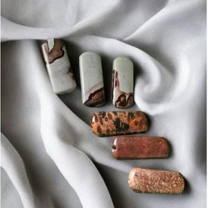 Peut inclure: Un assortiment de perles de pierres précieuses rectangulaires aux couleurs et motifs variés. Les perles présentent des nuances de blanc, de marron, d'orange et de cuivre, avec des veinures et textures uniques. Certaines perles sont percées.