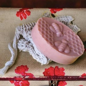 May include: A pink bar of soap with a honeycomb pattern and two bee designs, resting on a white washcloth. The soap is on a floral patterned surface with red flowers.