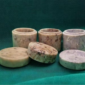 Pode incluir: Uma coleção de seis recipientes e tampas de pedra. Os recipientes cilíndricos e as tampas redondas são em tons de bege, cinza e marrom, com padrões salpicados. Os itens estão dispostos sobre um tecido verde escuro.