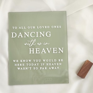 May include: A rectangular sage green acrylic sign featuring white raised lettering that reads: TO ALL OUR LOVED ONES DANCING with us in HEAVEN WE KNOW YOU WOULD BE HERE TODAY IF HEAVEN WASN'T SO FAR AWAY. A small rectangular wooden block stand is included.