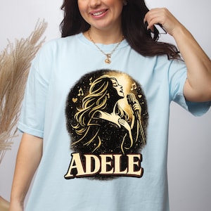 May include: Light blue t-shirt featuring a graphic of a singer with long hair holding a microphone, set against a black and gold background. The name "ADELE" is printed in a bold, stylized font below the image. Musical notes and hearts are also included.