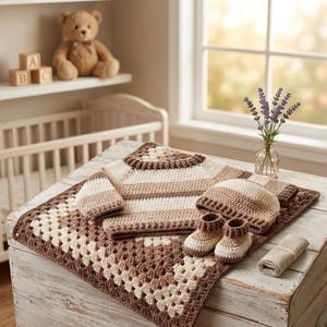 Può includere: Un set per neonati all'uncinetto composto da un maglione a maniche lunghe, un berretto e scarpine, tutti realizzati con strisce orizzontali color crema, marrone chiaro e marrone scuro. Il set è disposto su una coperta quadrata granny square.