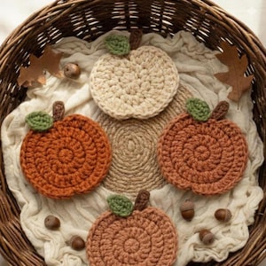 Peut inclure: Un panier en osier contient quatre dessous de verre en forme de citrouille crochetées, dans des tons orange et crème, chacune avec une feuille verte et une tige marron. Les dessous de verre reposent sur un tissu crème, avec des glands et des feuilles d'automne.